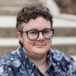 Headshot of Darcie Adams, a man with short brown curly hair and blue patterned glasses wearing a purple floral shirt.
